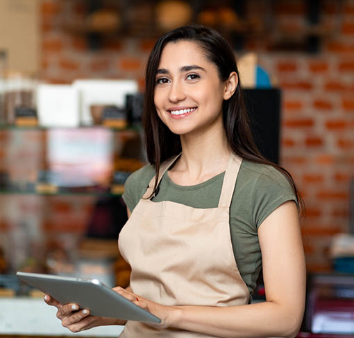 Opening small business. Happy arab woman in apron near bar counter holding digital tablet and looking at camera, waiting for clients in modern loft cafe