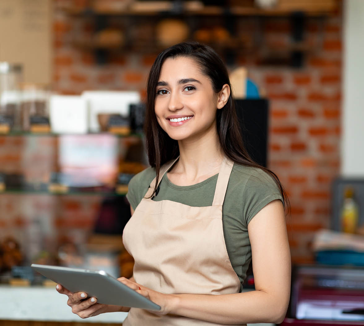 Opening small business. Happy arab woman in apron near bar counter holding digital tablet and looking at camera, waiting for clients in modern loft cafe